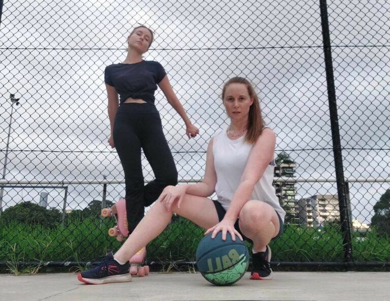 Carley Johnston is crouched on a basketball court with her left hand resting on top of a basketball. Her right hand is resting on her knee with her right leg outstretched. Carley is wearing a white tank top and black shorts with sneakers. Carley's friend is behind her leaning against the chain link fence. She is wearing a black crop top, long black pants and pink roller skates. Her head is tilted to the side and her left leg raised at a right angle.