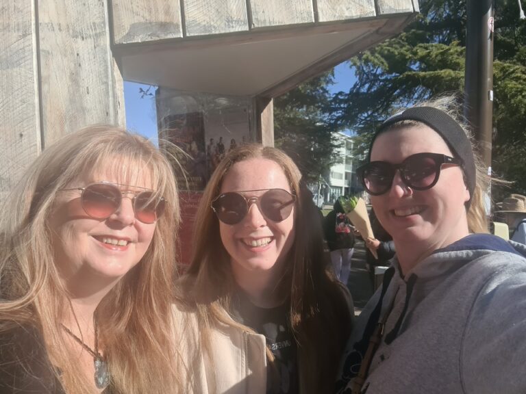 Carley Johnston is standing in the sunshine, between her mum and sister. Carley's wearing round sunglasses, a black jumper and cream coat. Carley's Sister is wearing a black headband, black rimmed sunglasses and a grey hoodie. Carley's mum is wearing Gold and bronze sunglasses a necklace made of leather with a blue glass heart in the centre and a black top.