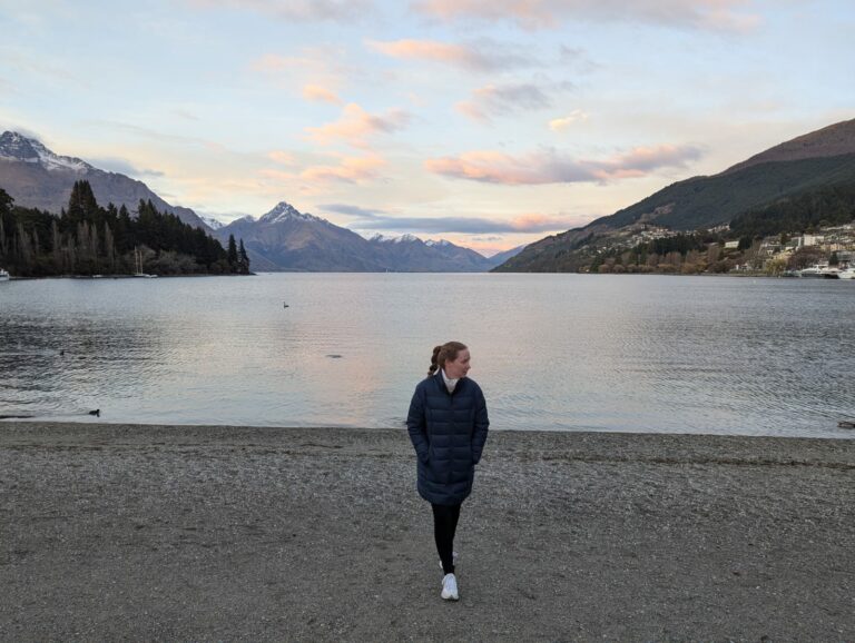 Carley Johnston stands at the edge of Lake Wakatipu in Queenstown, New Zealand. She is wearing a navy blue puffer jacket, black jeans, white sneakers and a pink scarf.